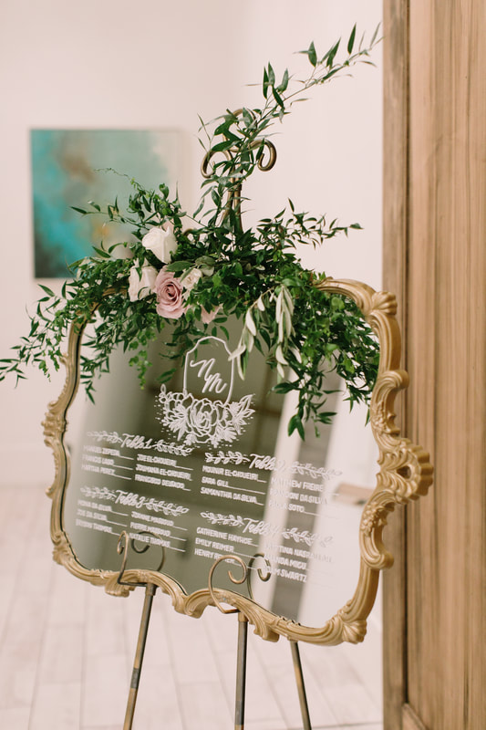 Gold mirror seating chart with modern calligraphy and beautiful, rustic floral decor on gold stand. Photo by Mangos Studio, Ontario, Canada. Featured in Carats. & Cake January 2022.